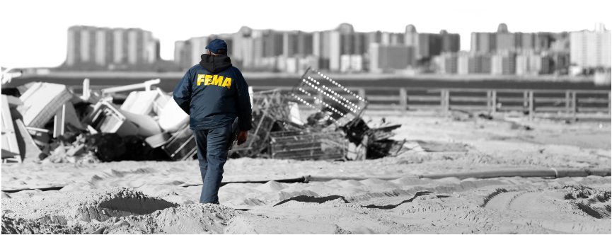 An inspector wearing a FEMA jacket walks among debris at a disaster site