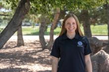 a photo of a woman with brunette hair standing with a dark blue FEMA polo shirt and standing in front of trees and rocks