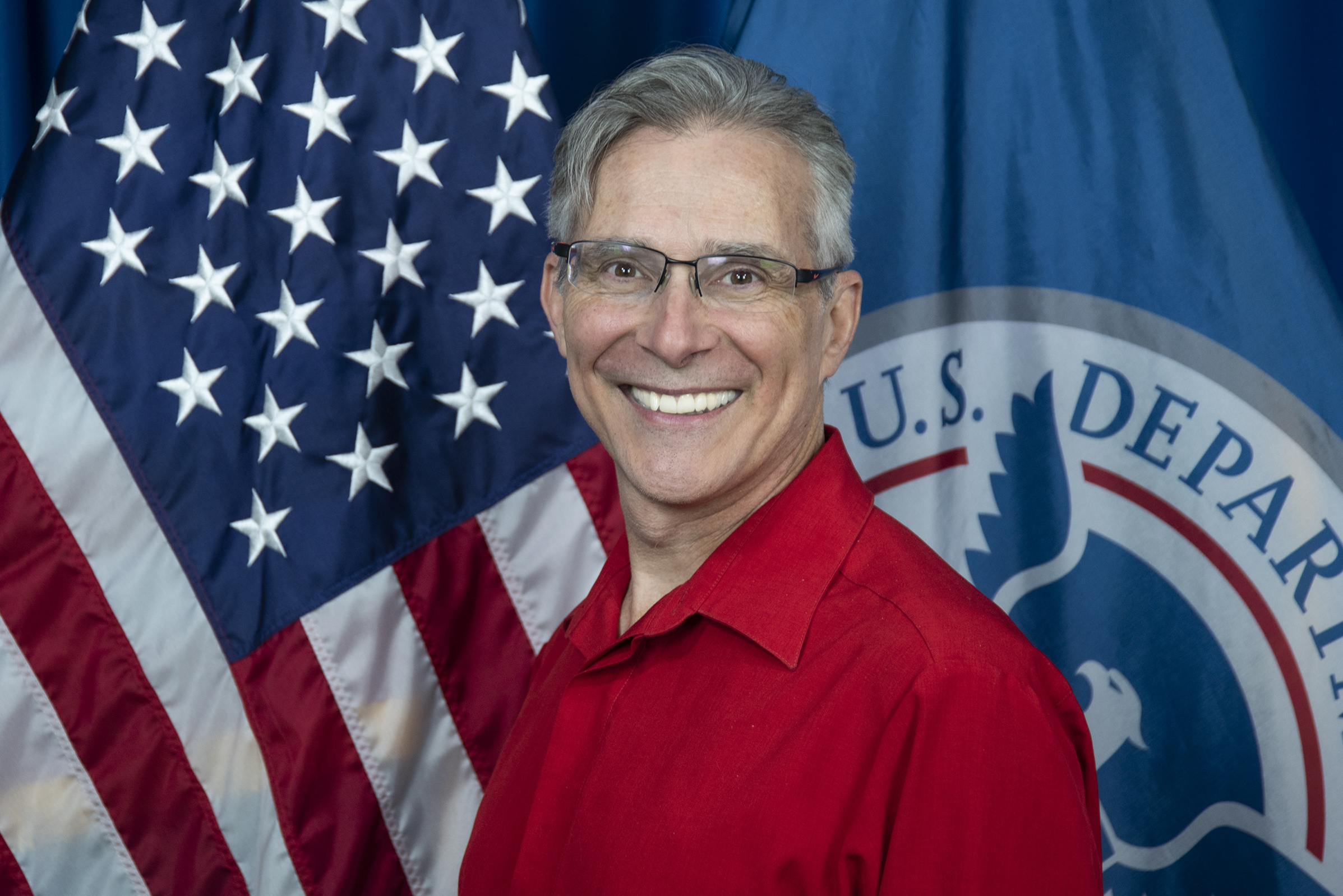 A photo of a man in a red shirt smiling and wearing glasses, with the background of the photo the United States flag on the left and the Department of Homeland Security flag on the right.