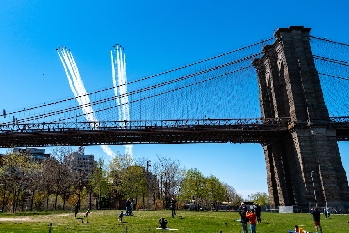 Jets fly over the Brooklyn Bridge as people watch from the ground.