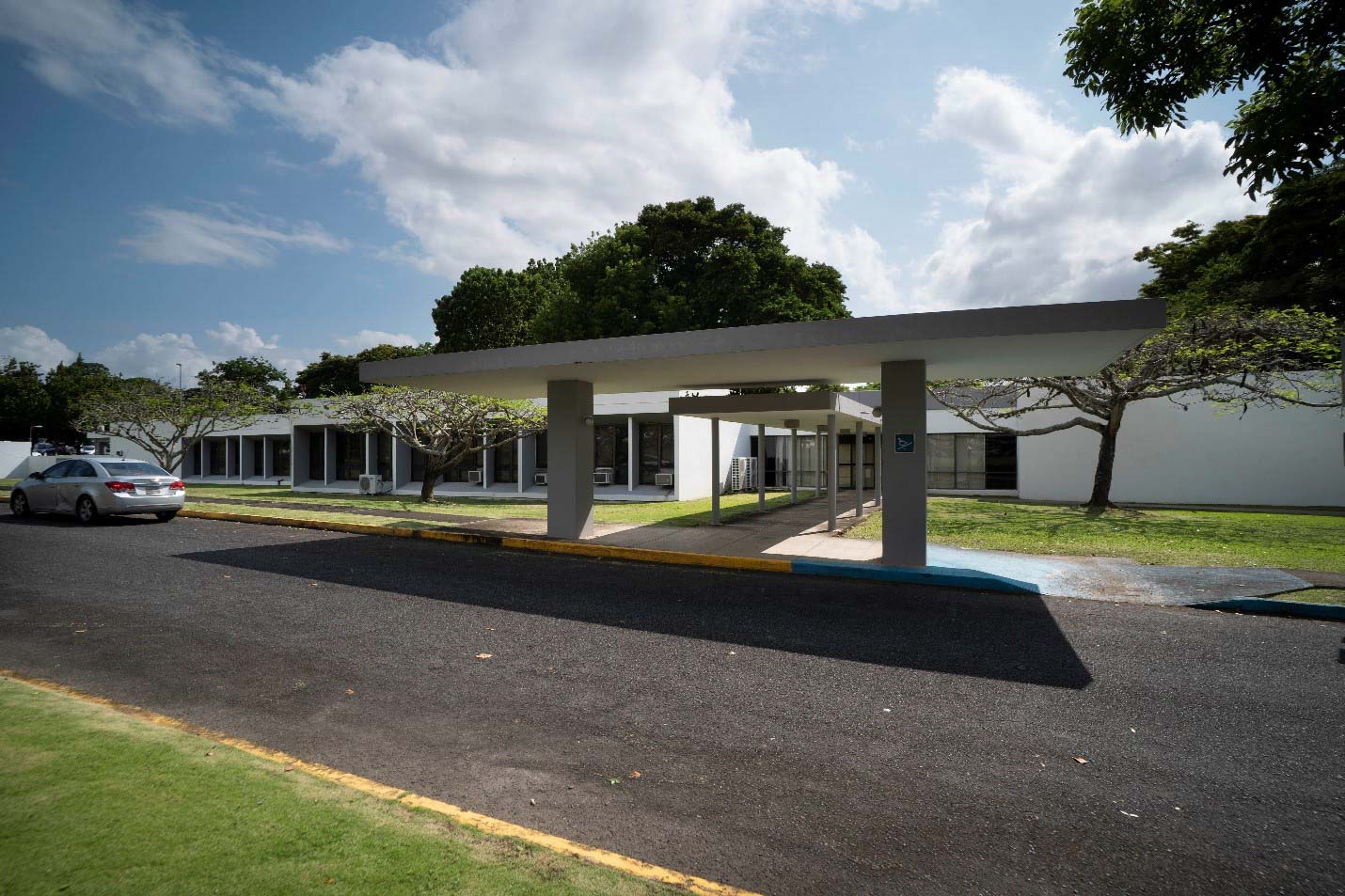 View of the entrance and building of the Central University of the Caribbean. One story white building with columns in front. One silver car parked to the left of the building.