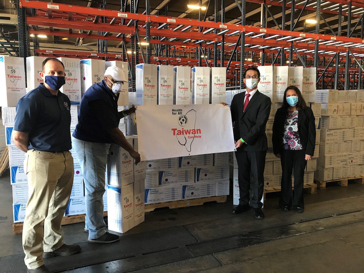 Four people wearing PPE masks holding up a sign in front of boxes of PPEs.