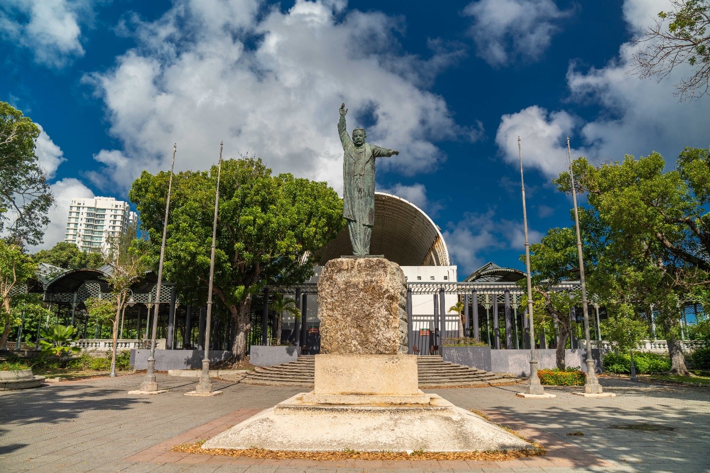 View of a statue of a man with hand raised. Trees and buildings on the background.
