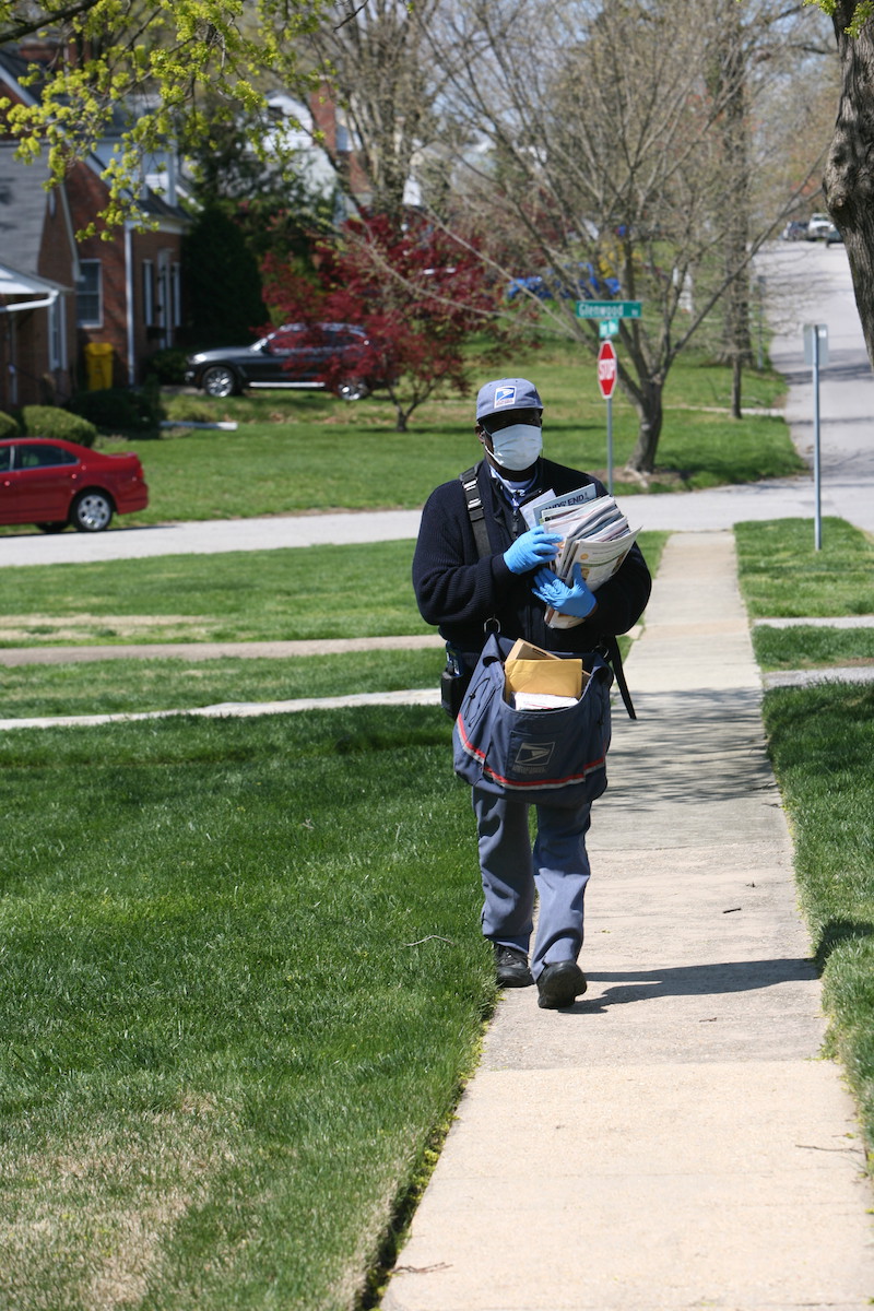 A postal service worker walking down the street carrying mail, wearing protective gloves and a mask 