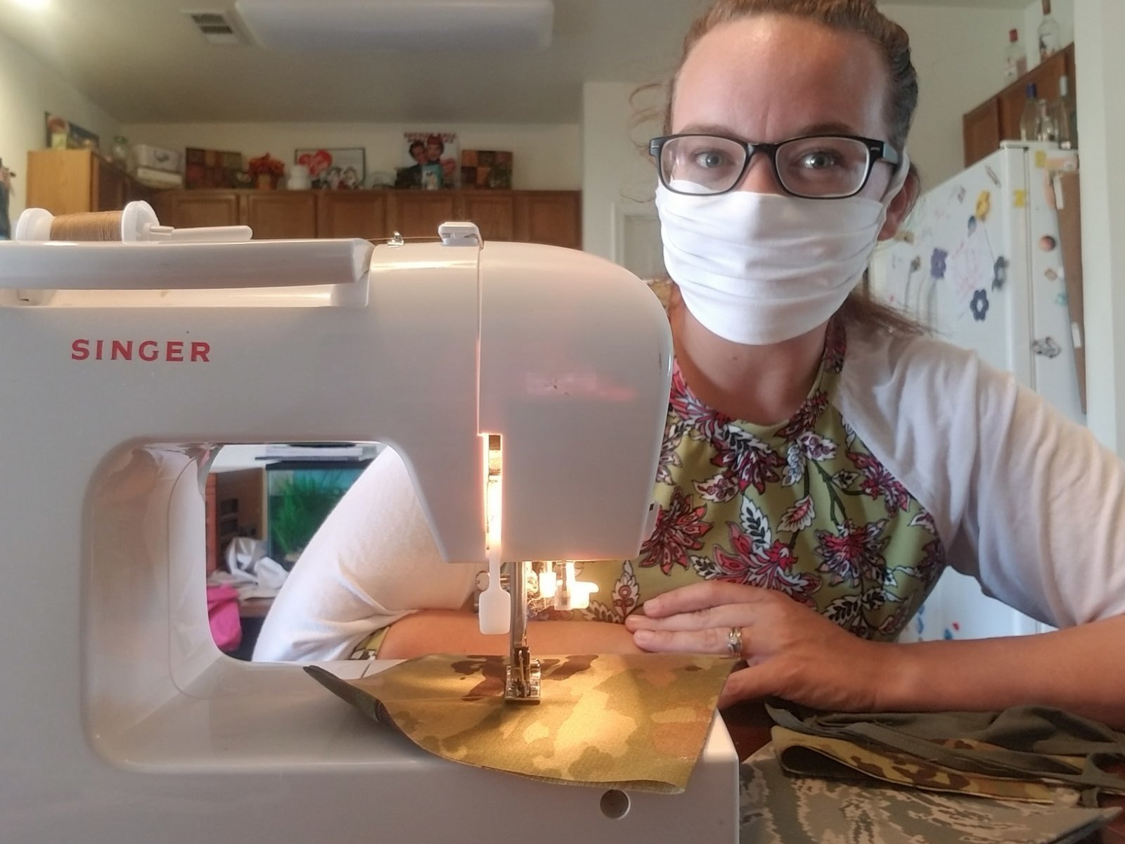 A woman in PPE mask looks into the camera and is sitting by a sewing machine.
