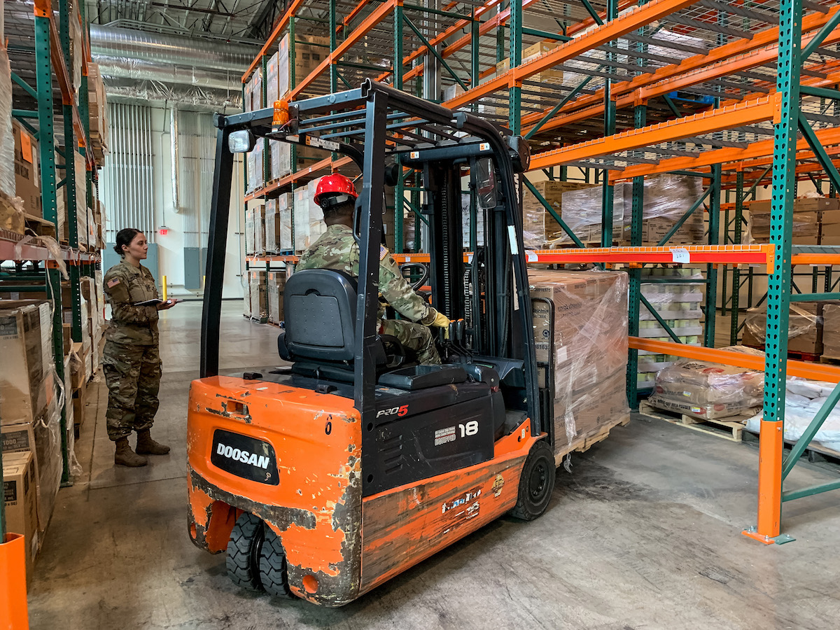 A forklift backs up from shelves with medical supplies as a standing solder conducts inventory.