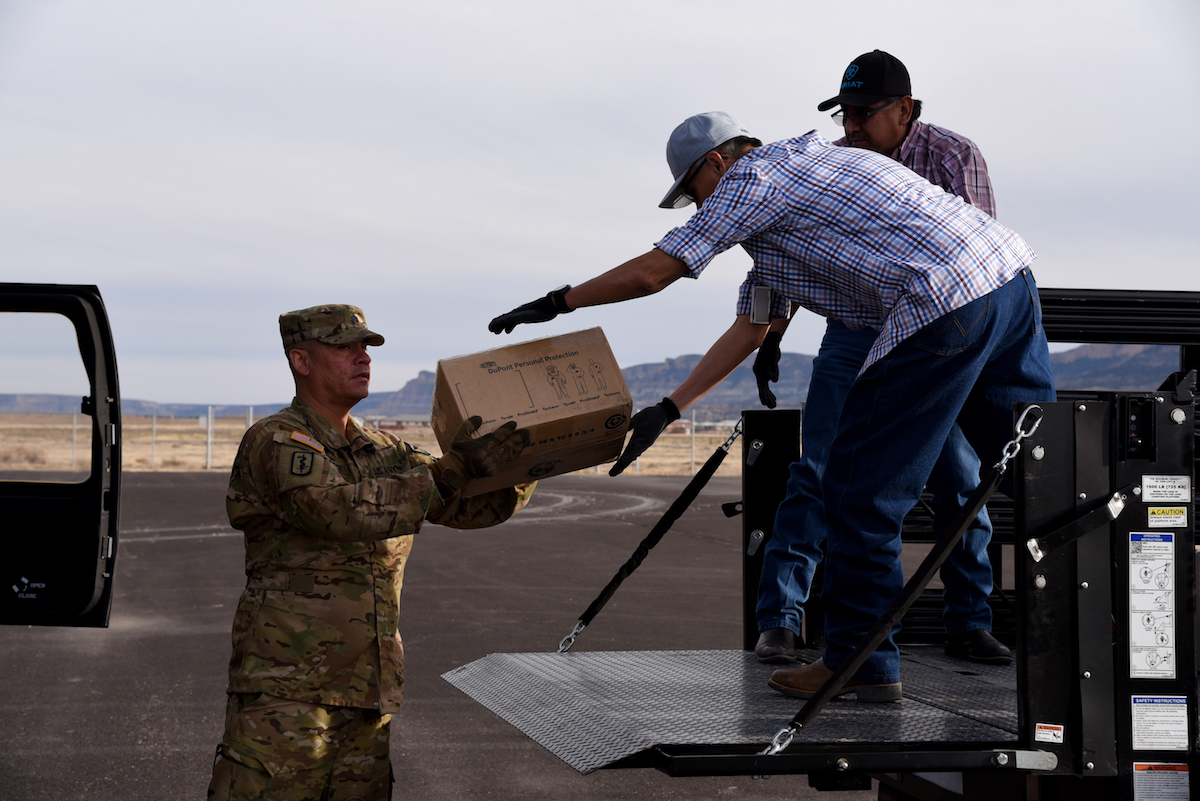 Soldier standing on the ground hands box of supplies to civilians in the back of a truck.
