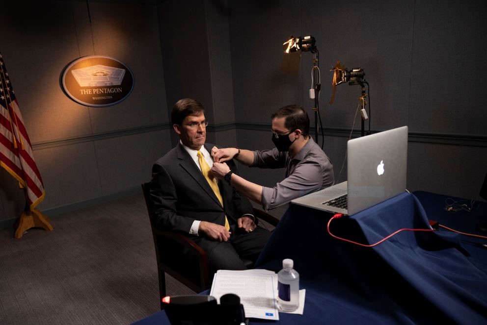 A male fixing Secretary of Defense Mark T. Esper's mic on him before he does a video call interview