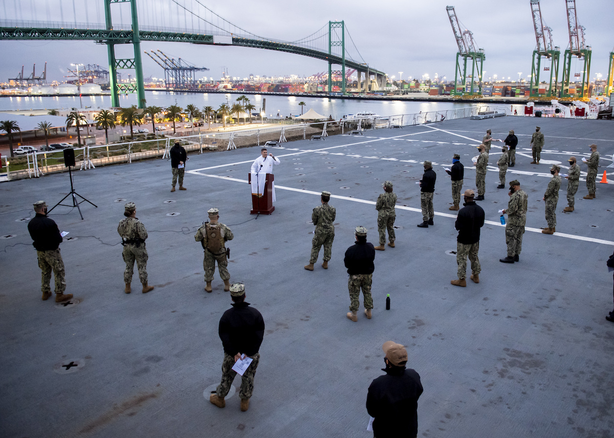 Military personnel stand spaced apart outside while listening to an Easter sermon.