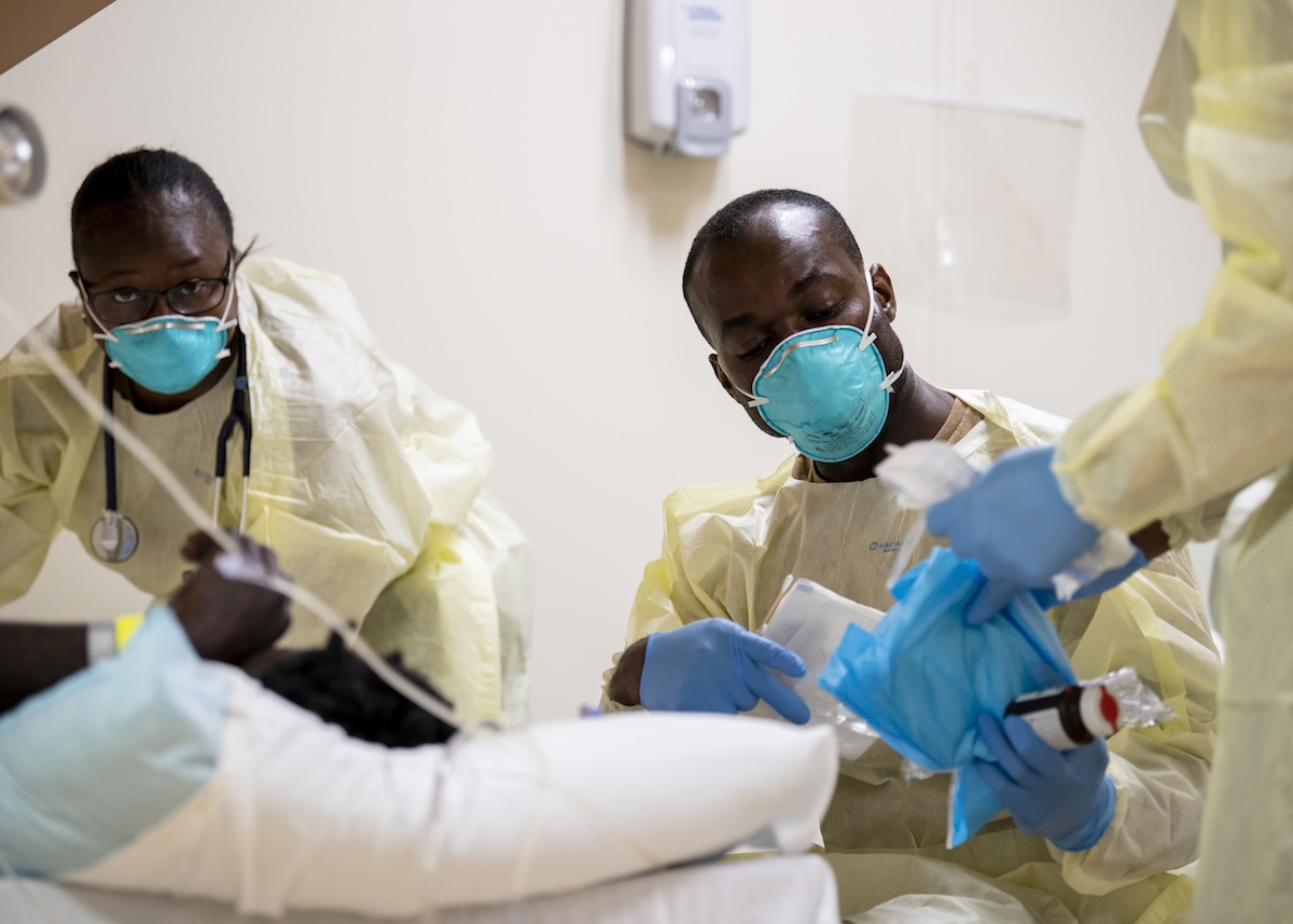Medical personnel in masks and gowns pass bloodwork and supplies to a third person. 