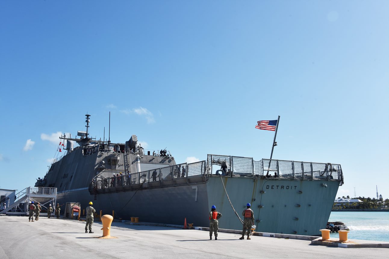 A US Navy ship with an American Flag on top with 6 soldiers standing outside of it.