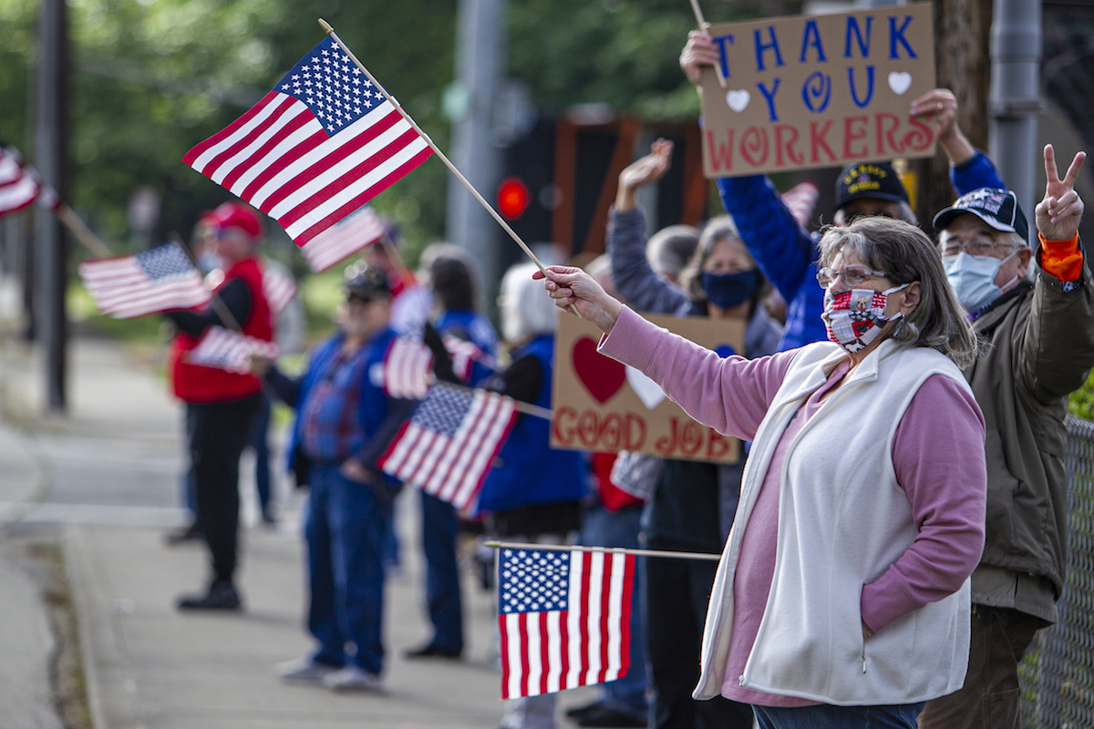 People in PPE masks waves American flags and Thank You signs on the side of a street.