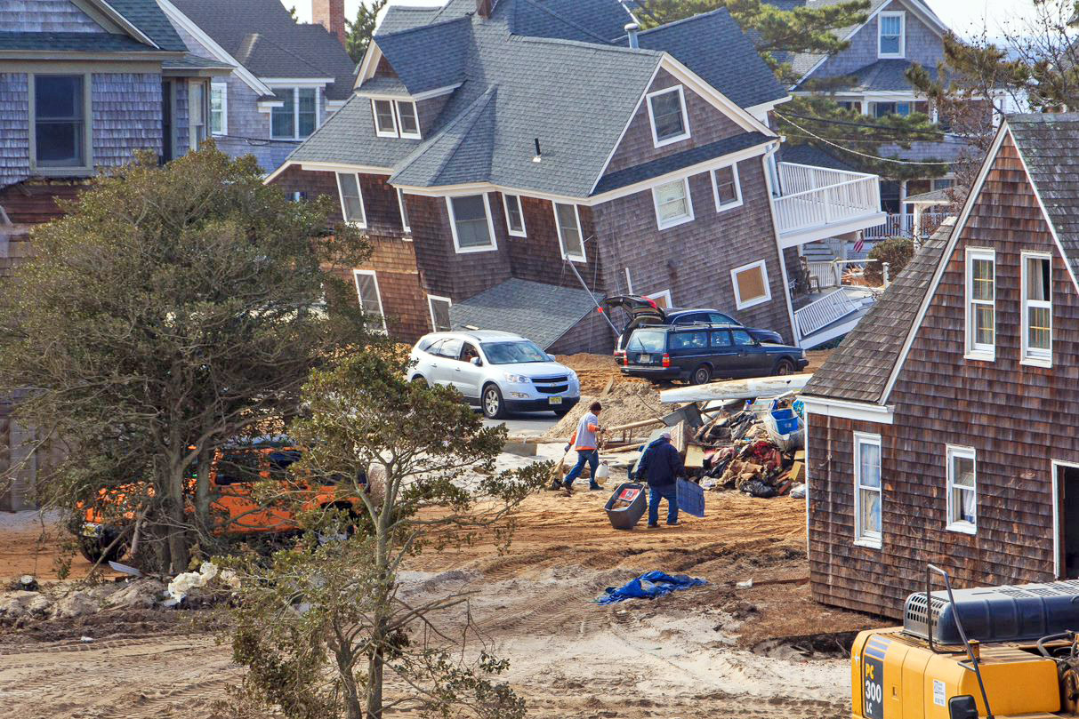 House buried in sand, two men clean up piles of debris. 