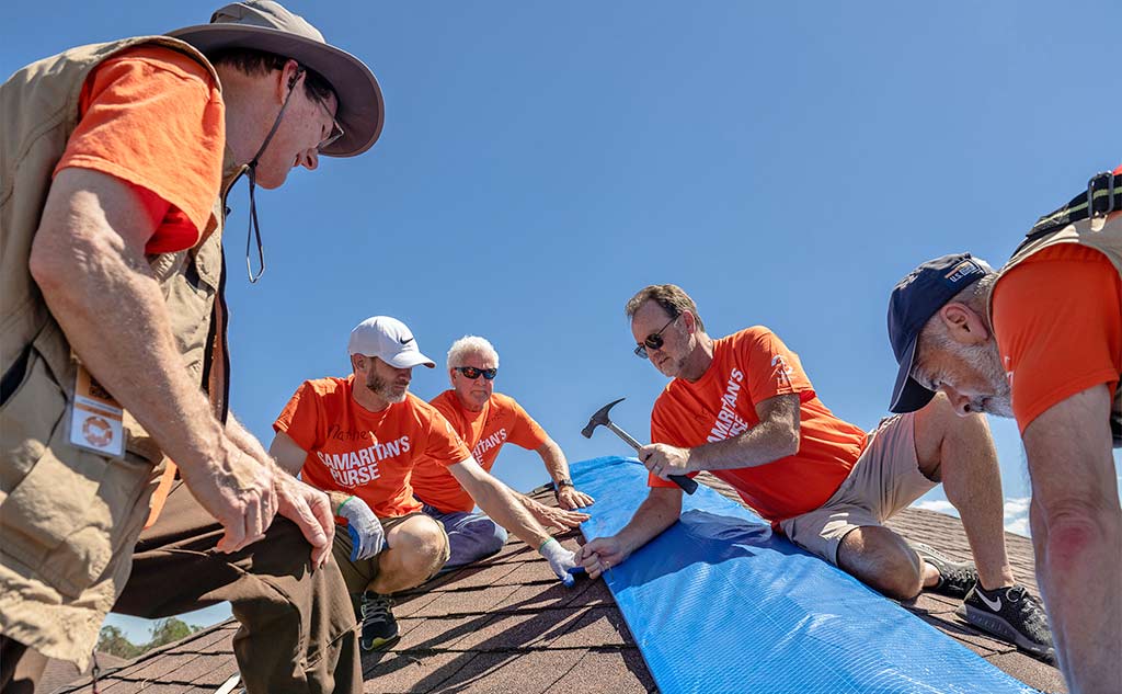 Five men sit and stand on roof to put down a blue tarp.