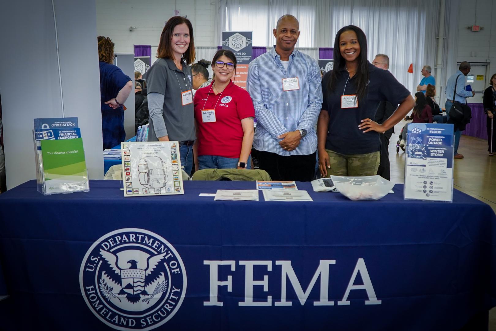 Four recruitment specialists stand smiling in front of FEMA table.