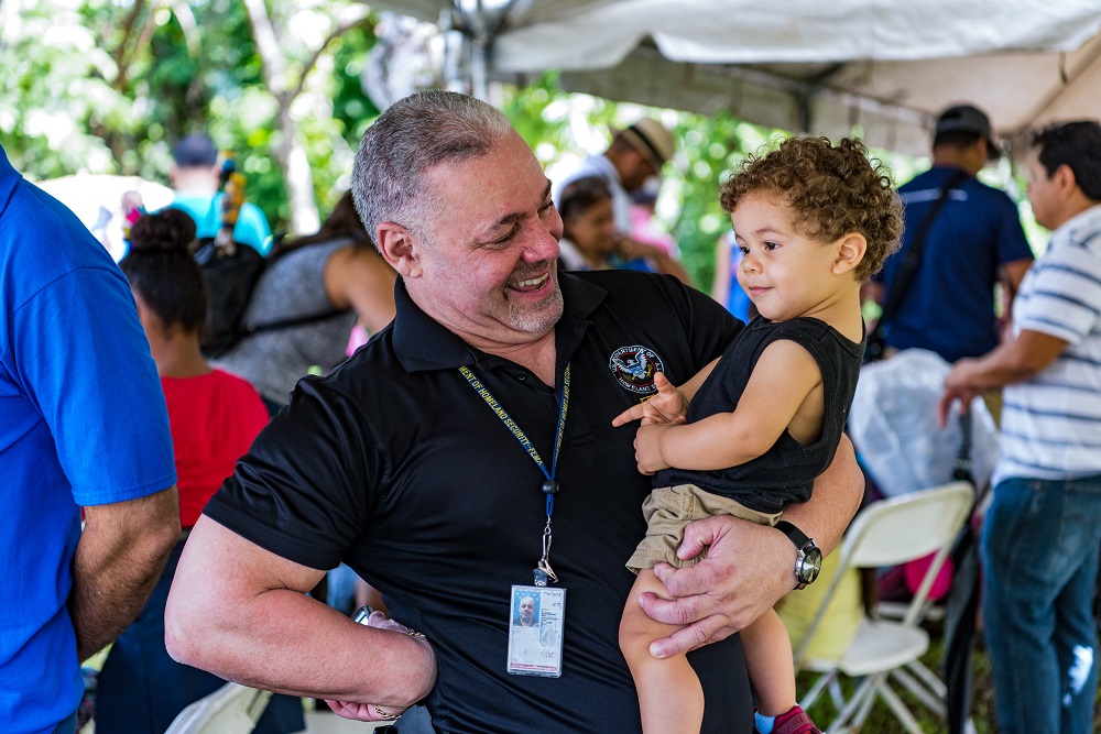 A man in FEMA polo shirt beams at a toddler he's holding in his arms.