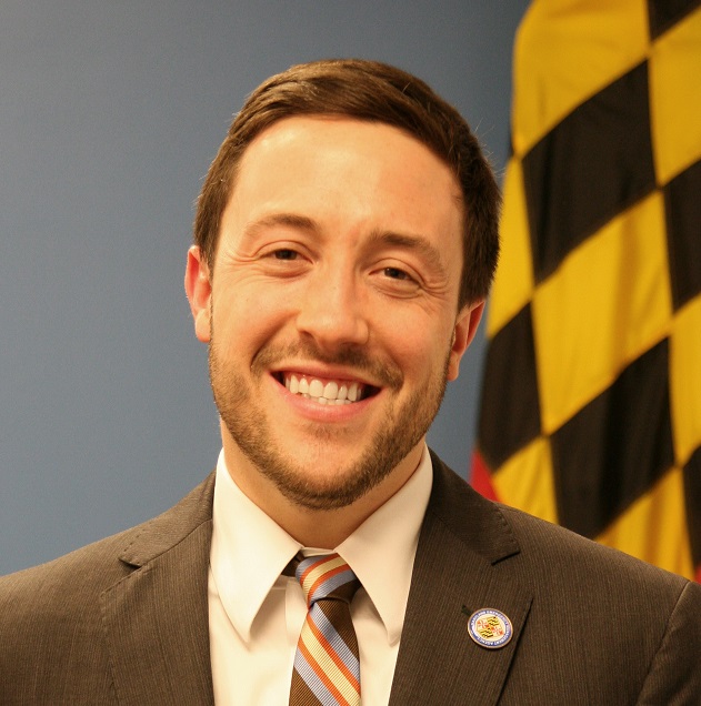 Man in suit stands in front of Maryland flag.