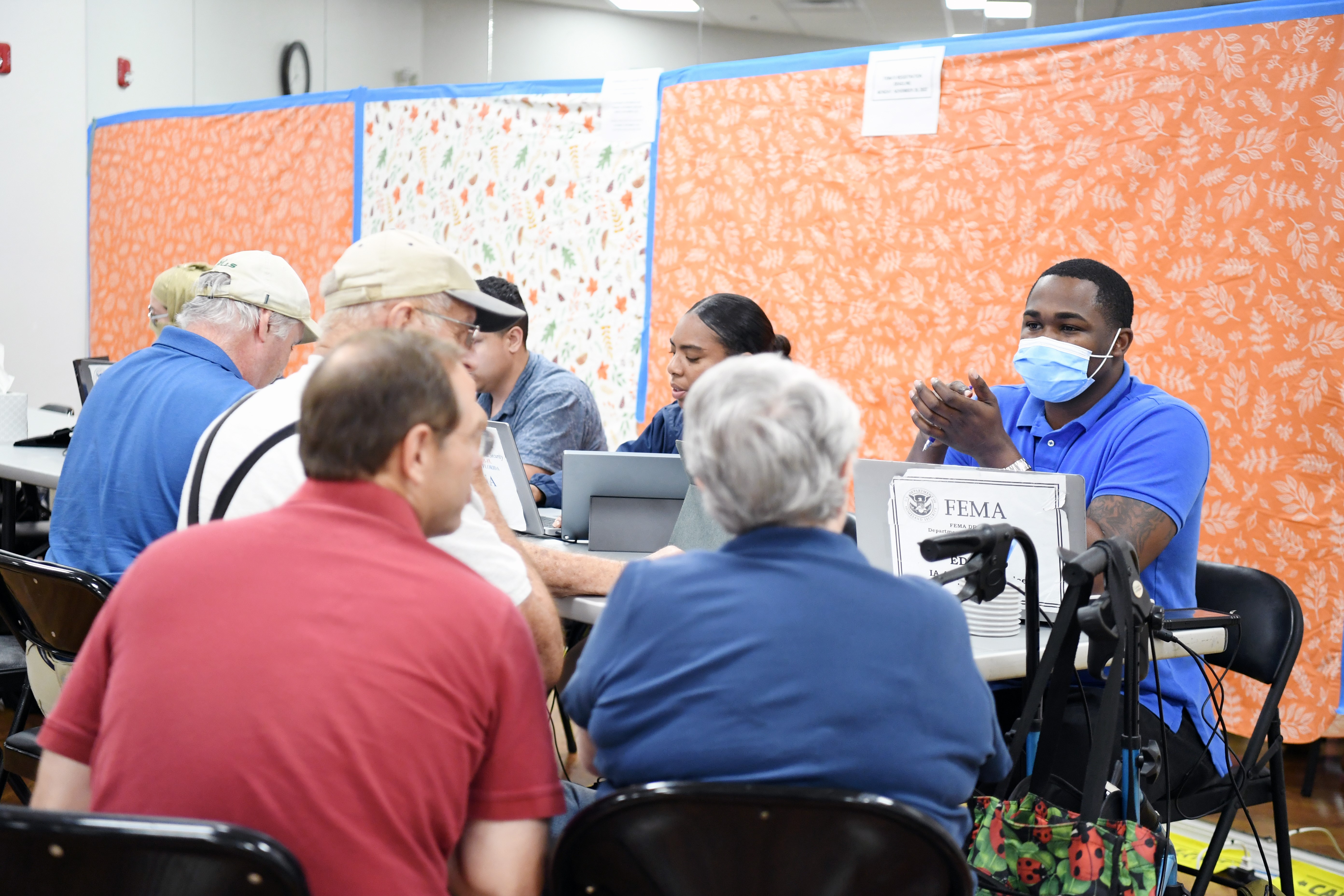 2 male & 1 female FEMA specialist at a table with their laptops talking to survivors on the other side or the table