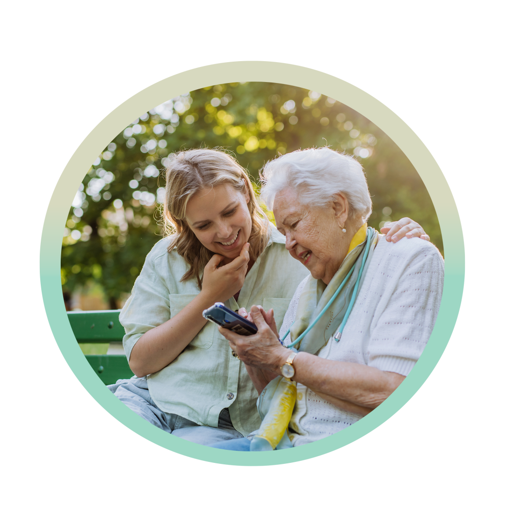 A young white woman sitting beside her white grandmother looking into a phone. They are outside on a sunny day on a bench. 