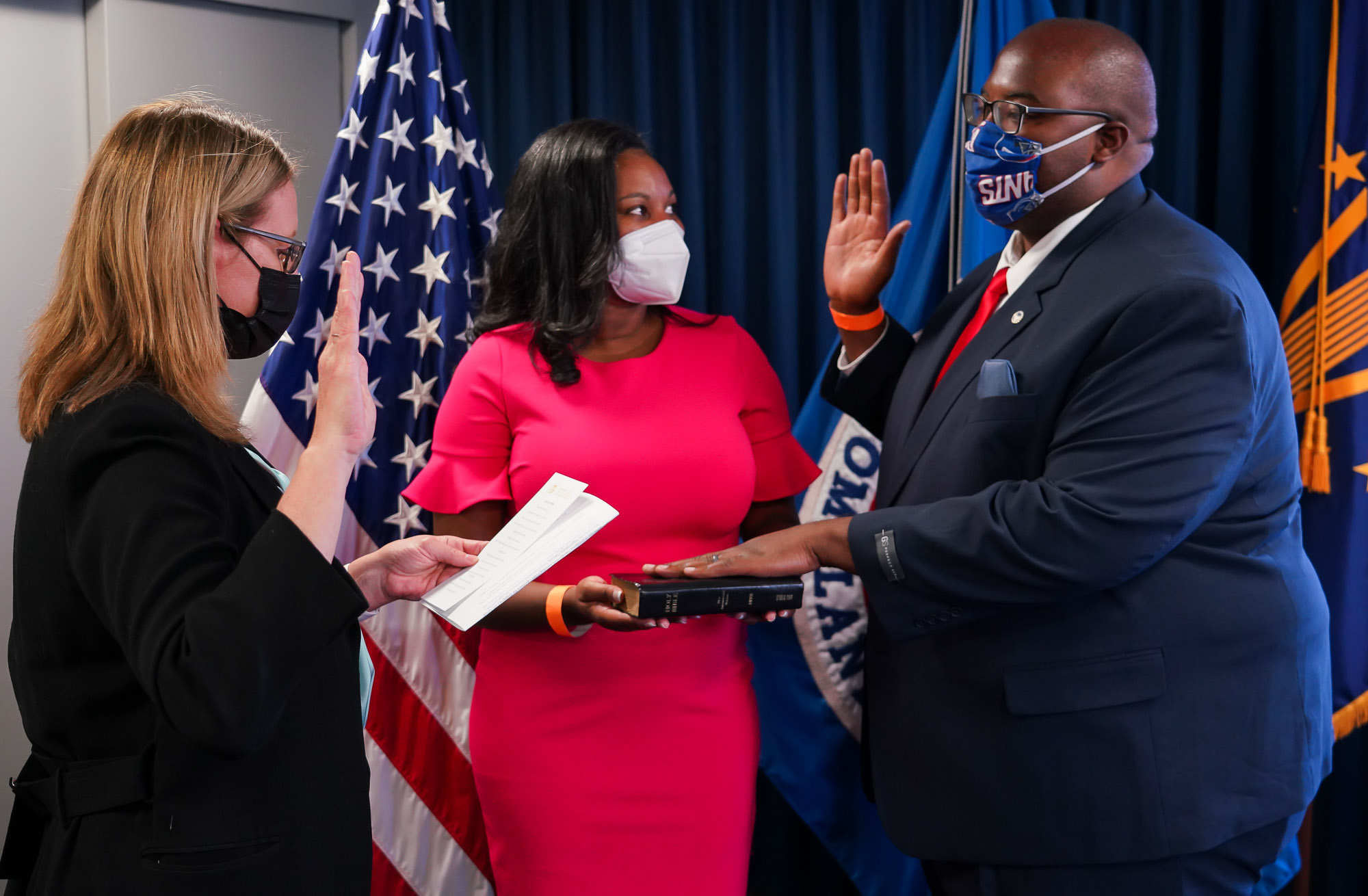 FEMA Administrator hold her hand up swearing Marcus in while his hand is on the bible that a woman is holding in the middle of them. 