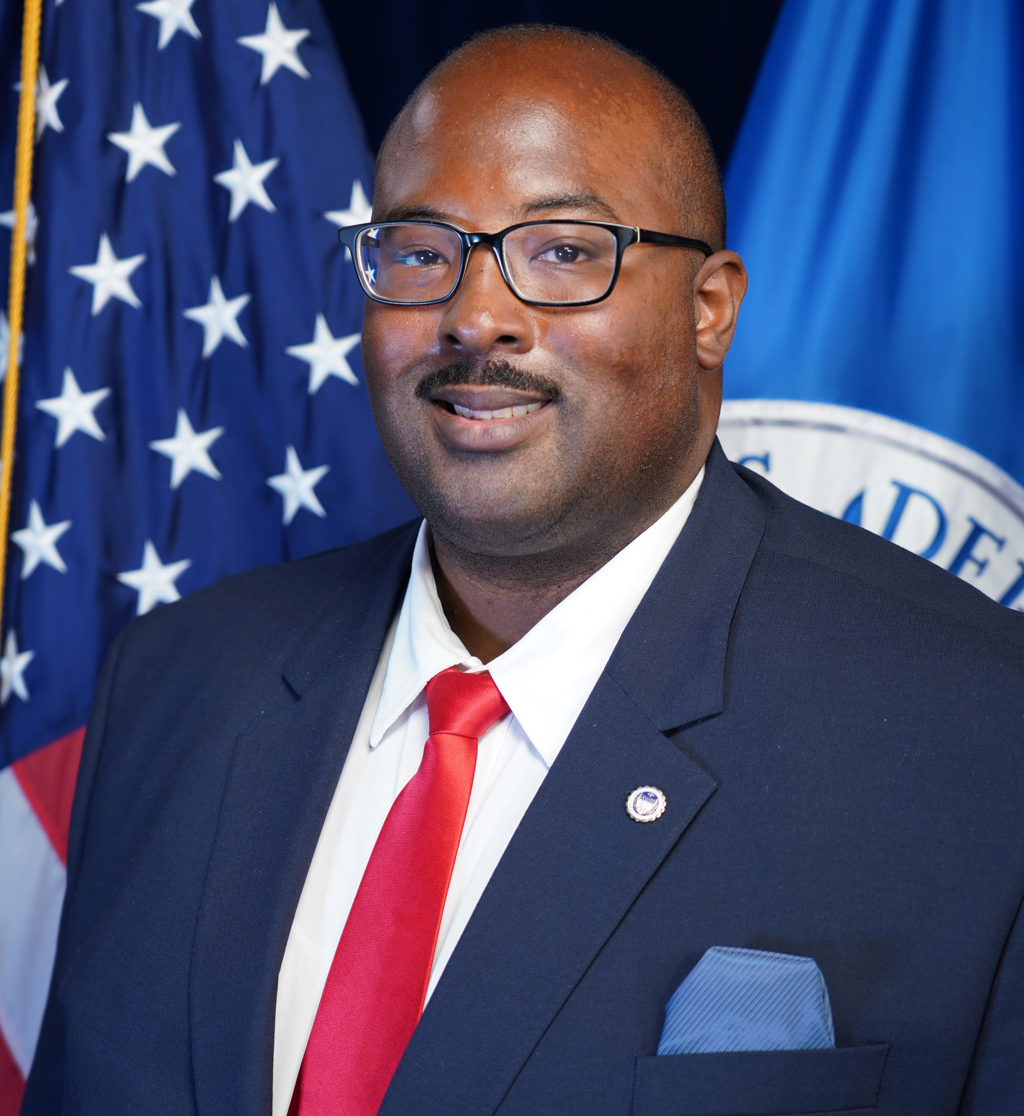 A African American Make with a blue suit on, red tie, white shirt and glasses while standing in front of the American flag and DHS flag