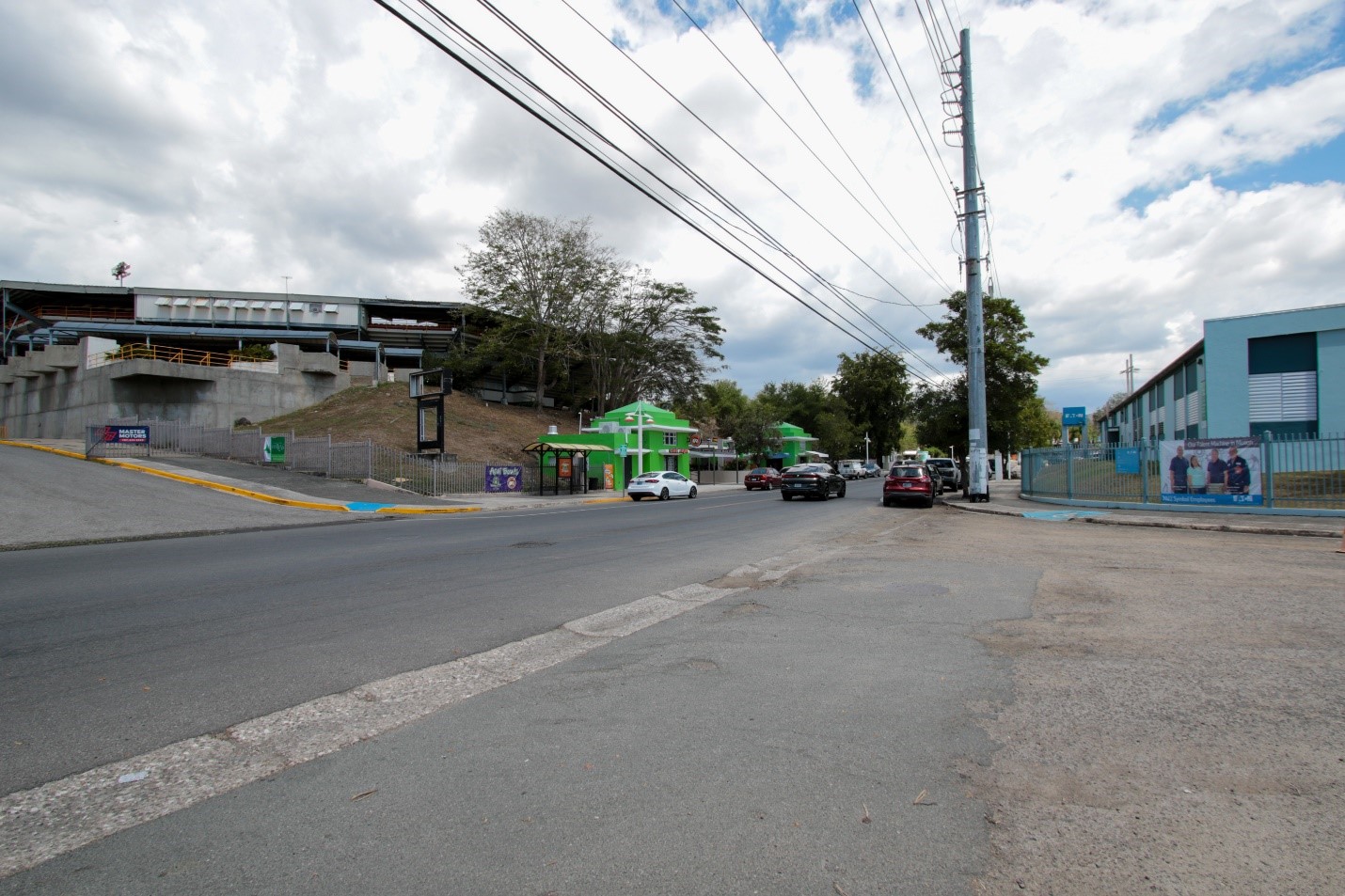 View of a road with green buildings to the left and blue and white building to the right. Several cars are transiting the area.  