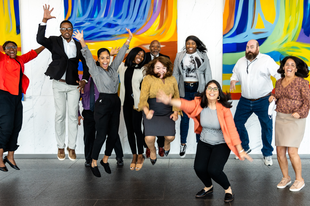 Ten adults in business casual clothes jumping in the air and laughing in front of brightly colored art.