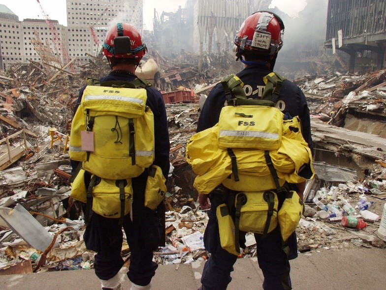 Two rescue workers wearing yellow packs and red helmets in front of debris