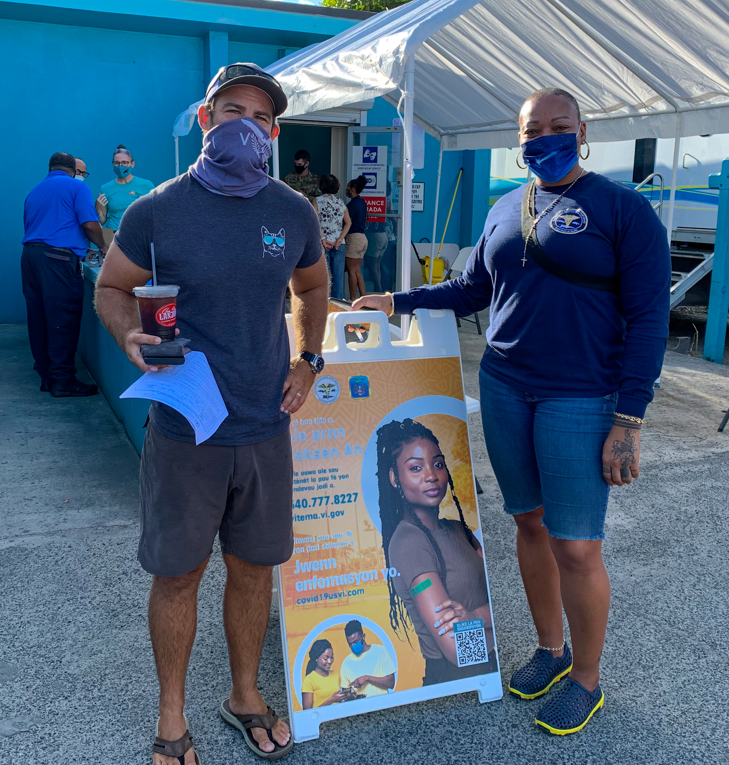 A male sanding on one side and a woman standing on the other side of a sign. They are are both wearing face mask. 