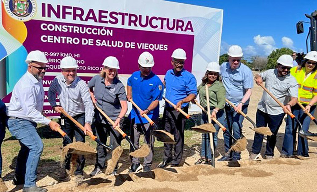 A row of people in hard hats holding shovels to the ground.