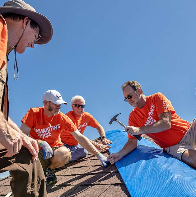 Men in matching orange shirts hammer a tarp to a roof.