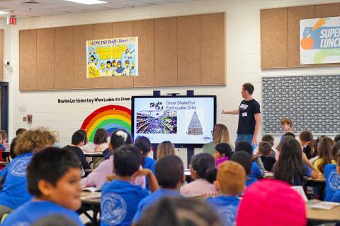 Caption: <p>Man points to large poster in front of an assembly of kids. </p>