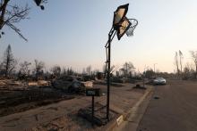 A basketball hoop stands twisted by heat. A car destroyed by fire sits in the driveway. 