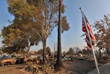 A US Flag stands in the foreground surrounded by a burned out vehicle and debris left from a fire. 