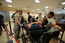  A man reclines on a portable hospital bed as the man at his feet tightens the straps holding his legs. A woman stands on his right side holding an IV bag. 