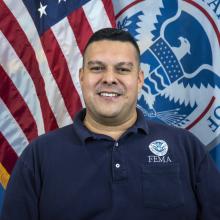 A man smiling in front of the American and DHS flag