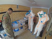 Two people putting on Personal Protective Equipment (PPE) with one soldier standing in front of them looking at a paper S