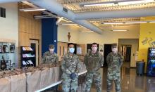 Military personnel stand around in PPE by tables that have bags full of food.