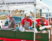 Three men stand at table under canopy serving food.