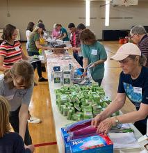 Group of people stand around table with supplies on it.